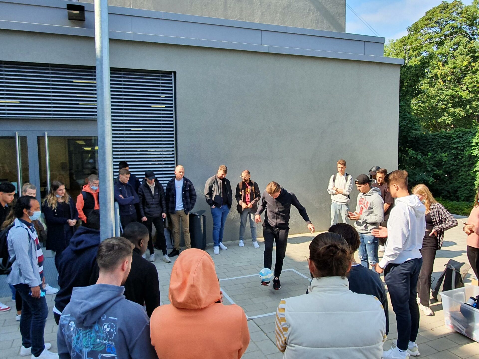 Gruppe von Berufsschülerinnen und Schülern stehen um einen Schüler herum, der einen Fußball auf seinem Spann balanciert.