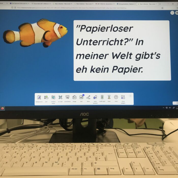 Ein Monitor. Darauf ein Bild von einem bunten Fisch. Dieser sagt: „Papierloser Unterricht? In meiner Welt gibt's eh kein Papier.“