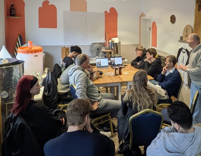 Zu sehen ist eine Gruppe Menschen in einem Raum, die alle auf einen Laptop schauen, auf dem eine Präsentation gezeigt wird. Die Personen werden von hinten gezeigt, einige sitzen um einen Tisch herum, andere dahinter auf Stühlen.
